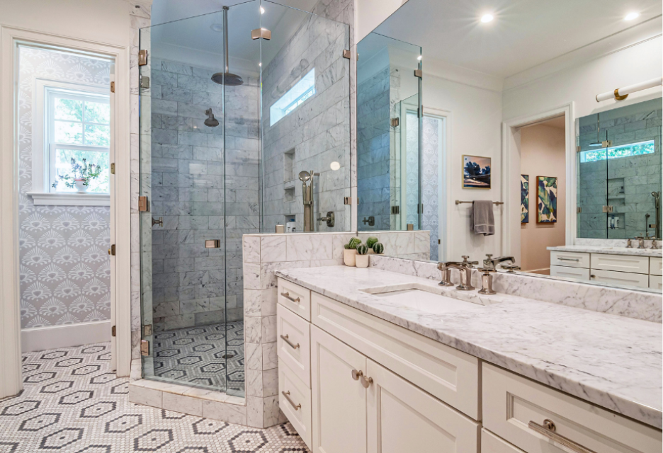 large marble bathroom with a long counter top on the right.  Countertop is light baige.  ther is a large corner standing show next to the counter and an open doorway to a small close to the left of it. 